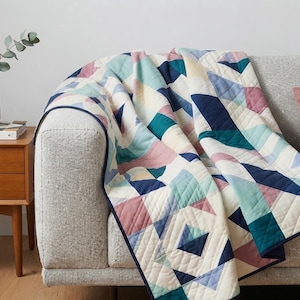 May include: A quilted throw blanket featuring a geometric pattern of triangles and diamonds in shades of navy blue, teal, light blue, cream, and dusty rose. The blanket has a dark blue fabric border and visible diagonal stitching throughout.