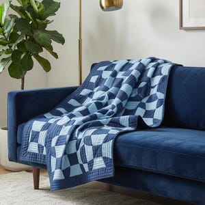 May include: A quilted throw blanket featuring a geometric pattern of alternating dark navy blue and light blue curved shapes. The blanket has a stitched grid texture throughout and is draped over a solid dark navy blue velvet sofa.