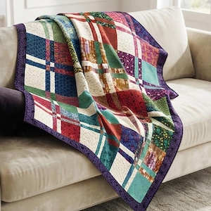 Puede incluir: Una colcha de retazos con una cuadrícula de cuadrados de tela multicolores en tonos rojo, azul, verde, morado y crema. La colcha está terminada con un borde morado oscuro que presenta un patrón geométrico repetitivo y costuras.