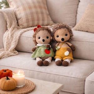 May include: Two handmade crochet hedgehog dolls sit on a beige sofa. One wears a green dress with a red apple, the other wears yellow overalls. A small table holds pumpkins and a lit candle.