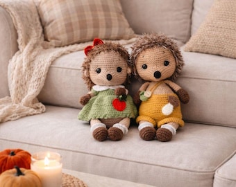 May include: Two handmade crochet hedgehog dolls sit on a beige sofa. One wears a green dress with a red apple, the other wears yellow overalls. A small table holds pumpkins and a lit candle.