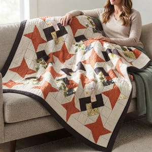 May include: A quilt featuring a repeating star pattern in burnt orange, dark charcoal, and cream with floral accents. The quilt has a dark charcoal border and a grid-like stitched texture throughout the fabric surface, creating a geometric design.