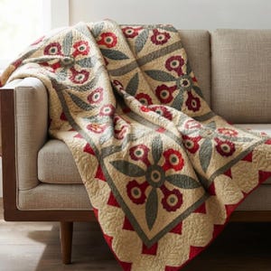 May include: A cream-colored quilt featuring a repeating pattern of dark red floral motifs and muted green leaf shapes. The fabric has a visible quilted texture throughout, with a scalloped red border edge that frames the central geometric design.