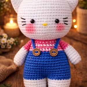 Puede incluir: Muñeca de gato de ganchillo hecha a mano con cabeza blanca, mejillas rosas y ojos negros. Viste un peto azul con botones de madera y una camiseta a rayas rosas y blancas. Sobre un fondo de madera cálido.