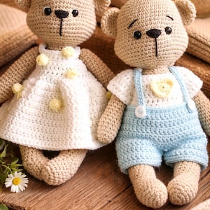 May include: Two handmade crochet teddy bears. One bear wears a white dress with yellow flower accents, the other wears blue overalls. Both are tan with black eyes and noses. The bears are sitting on a wooden surface.