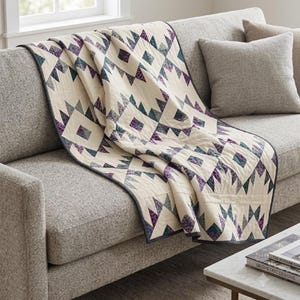 May include: A quilt featuring a geometric pattern of cream, purple, and teal triangles and diamonds. The fabric has a visible stitched texture throughout, and the entire piece is finished with a dark grey binding along the outer edges.