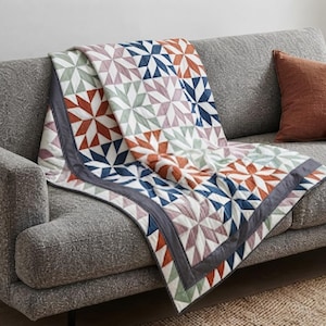 May include: A quilted throw blanket featuring a repeating star pattern in shades of rust orange, navy blue, sage green, and dusty rose. The quilt is finished with a solid dark gray border and displays visible stitching lines throughout the fabric.