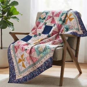 Puede incluir: Una colcha de patchwork con un patrón de molinillo que presenta varios cuadrados de tela floral y sólida en tonos rosa, azul, amarillo y verde. La colcha tiene un borde floral azul oscuro y está colocada sobre un sillón tapizado de color claro.