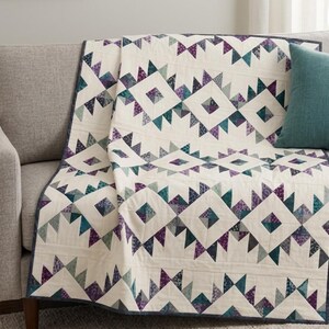 May include: A quilted blanket featuring a geometric pattern of diamonds and triangles in shades of purple, teal, and dark blue against a cream-colored background. The quilt has a dark blue border and is draped over a gray upholstered sofa.