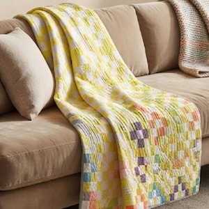 Può includere: Una trapunta patchwork caratterizzata da una griglia di piccoli quadrati nei toni del giallo, bianco, azzurro, arancione e viola. La trapunta presenta una visibile trama trapuntata e un bordo grigio chiaro, drappeggiata su un divano neutro.