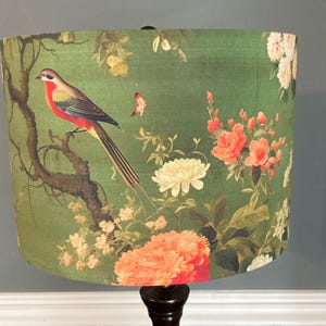 May include: A lampshade with a botanical design featuring a bird perched on a branch, surrounded by flowers and foliage. The color palette includes shades of green, red, and white. The lampshade is cylindrical and sits on a dark brown base.