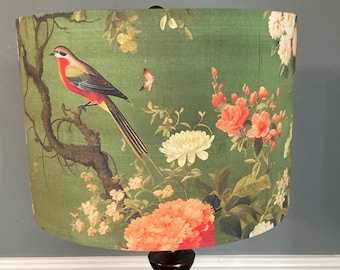 May include: A lampshade with a botanical design featuring a bird perched on a branch, surrounded by flowers and foliage. The color palette includes shades of green, red, and white. The lampshade is cylindrical and sits on a dark brown base.