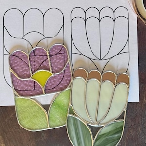 Puede incluir: Dos diseños de tulipanes de vidrieras, uno morado y amarillo, el otro crema y marrón, se encuentran sobre un papel blanco con un contorno de tulipán. El texto "SWEDA SISTERS" es visible. Una pequeña planta suculenta y cinta dorada están en el fondo.