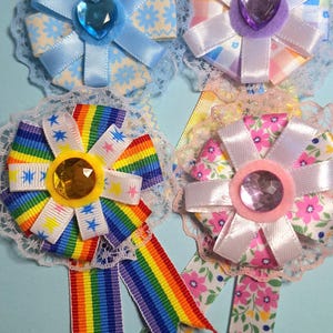May include: Four handmade ribbon rosettes with lace trim. The rosettes feature various designs, including floral patterns, rainbow stripes, and star accents. Each rosette has a central jewel and ribbon streamers, perfect for crafts or embellishments.