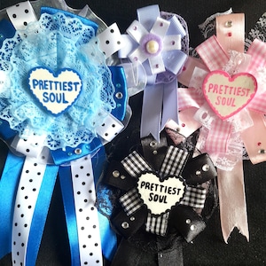 May include: Four decorative rosette badges with the text "Prettiest Soul" on heart-shaped centerpieces. The badges feature various colors, including blue, pink, black, and white, with ribbons, lace, and embellishments.