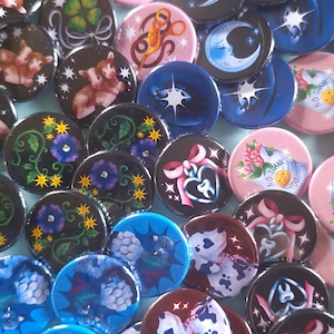 May include: A selection of circular badges with diverse designs. Some badges display animal illustrations, floral patterns, and celestial motifs. Others feature abstract designs and text. The badges are in shades of pink, blue, black, and brown.