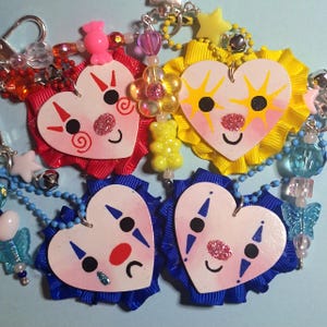 May include: A selection of handmade keyrings with heart-shaped clown faces, glitter noses, and ribbon trim. Each keyring has a unique colour scheme, including red, yellow, and blue, with various bead accents and charms.