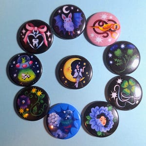 May include: A collection of ten circular pin-back buttons featuring various illustrations including a bat, a heart with a tooth, a UFO with a cow, a person on a crescent moon, a thistle, a four-leaf clover, a person in a blue flower, and a bird.