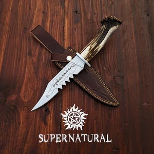 May include: A silver-toned metal knife with a serrated edge and engraved script on the blade, featuring a textured antler handle. The knife rests on a brown leather sheath with visible stitching. Below the items is a white Supernatural logo.