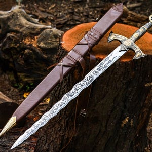 Puede incluir: Una espada de hoja ondulada con una empuñadura de metal ornamentada con detalles dorados y una piedra oscura en el pomo. La hoja muestra intrincados patrones grabados en negro. Se acompaña de una vaina de cuero marrón con punta dorada.