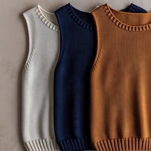 May include: Three sleeveless knit vests in a row, each with a ribbed neckline and hem. The colours are off-white, navy blue, and a warm brown. The vests are made of a textured knit fabric.