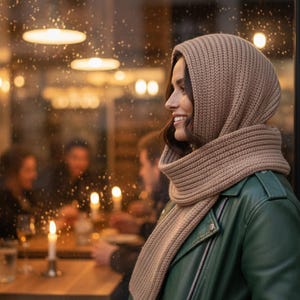 Könnte beinhalten: Eine Frau trägt ein beigefarbenes Strick-Hoodie- und Schal-Set, kombiniert mit einer grünen Lederjacke. Der Schal ist um den Hals gewickelt und die Kapuze bedeckt den Kopf. Der Hintergrund zeigt ein Restaurant mit sanfter Beleuchtung.