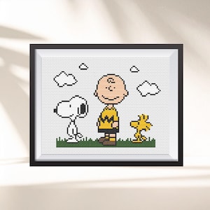 May include: Framed cross-stitch art featuring Snoopy, Charlie Brown, and Woodstock. The characters are depicted in a pixelated style, standing on green grass with white clouds in a blue sky. The artwork is framed in a black frame.
