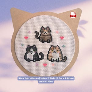 May include: A cross-stitch pattern featuring three cartoon cats in black, brown, and tan, with pink hearts and floral accents. The design is on a circular canvas, measuring 3.6 inches wide and 3.9 inches high, mounted on a cat-shaped cardboard backing.