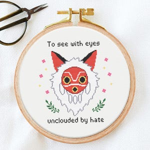 May include: A circular wooden embroidery hoop containing white fabric with a cross-stitch design of a red and white fox mask surrounded by small pink flowers and green leaves. The text reads To see with eyes unclouded by hate.
