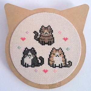 Peut inclure: Une broderie au point de croix représentant trois chats de dessin animé noirs, bruns et beiges, entourés de cœurs roses et de petites fleurs. Le motif est monté sur un support en carton en forme de chat.