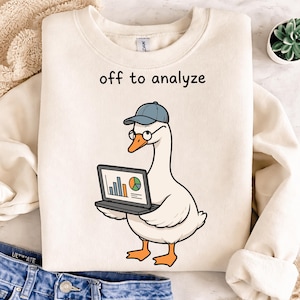 May include: Cream-colored sweatshirt with the phrase "off to analyze" printed at the top. A cartoon goose wearing a blue cap and glasses is holding a laptop displaying charts and graphs. The goose has orange feet and a beak.