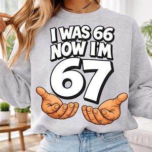 May include: A light gray sweatshirt with the text "I WAS 66 NOW I'M 67" in a bold, black font. The number "67" is large and white, with cartoon hands cupped below. A fun birthday gift.