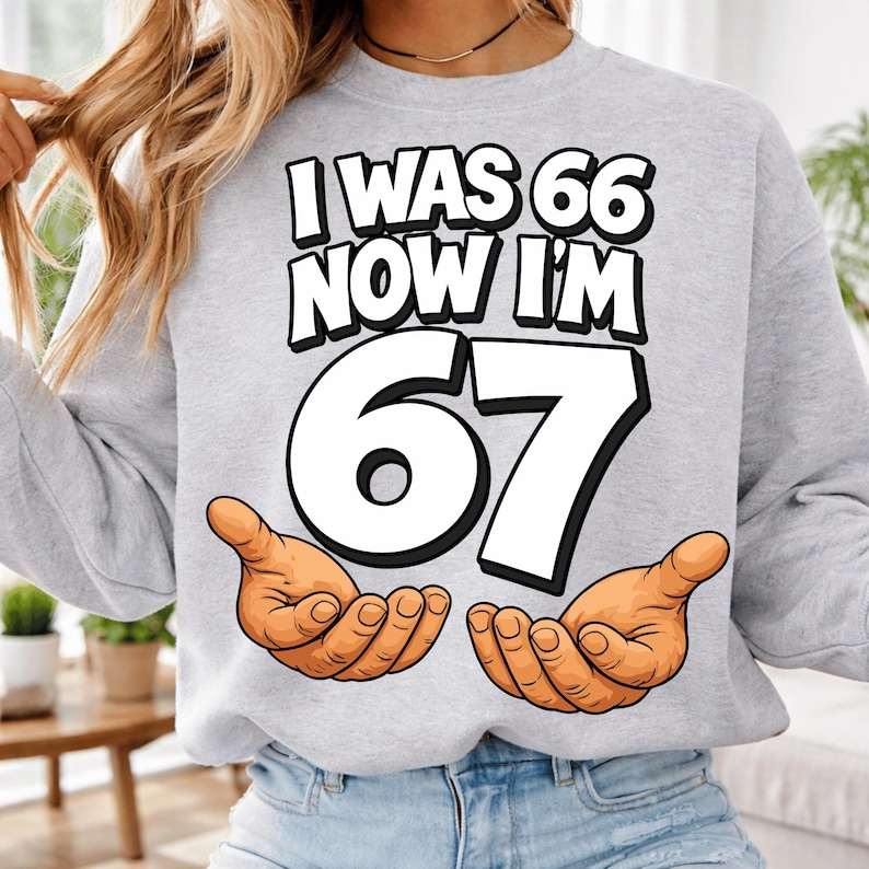May include: A light gray sweatshirt with the text "I WAS 66 NOW I'M 67" in a bold, black font. The number "67" is large and white, with cartoon hands cupped below. A fun birthday gift.