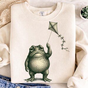 May include: Cream-colored sweatshirt featuring a detailed illustration of a frog holding a kite. The frog has a sad expression, and the kite is adorned with bows. The design is in a vintage, engraved style, creating a whimsical and artistic aesthetic.