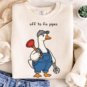 以下が含まれることがあります： 「off to fix pipes」の文字が入ったクリーム色のスウェットシャツ。グレーの帽子と青いオーバーオールを着た漫画のガチョウが、ラバーカップとレンチを持っています。ガチョウはオレンジ色のくちばしと足を持っています。
