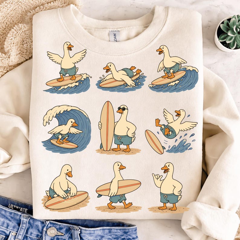Pode incluir: Uma camisola de gola redonda cor de creme com uma impress&atilde;o gr&aacute;fica de nove patos de desenhos animados em v&aacute;rias poses de surf. Os patos usam cal&ccedil;&otilde;es de banho azuis e interagem com pranchas de surf e ondas do mar azuis.