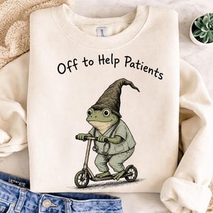 Puede incluir: Sudadera color crema con el texto "Off to Help Patients". El gráfico presenta una rana con uniforme médico y un gorro puntiagudo, montada en un patinete. Un estetoscopio cuelga del cuello de la rana.
