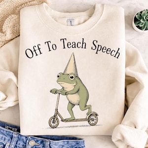 Op de afbeelding: Crèmekleurige sweatshirt met de tekst "Off To Teach Speech". De afbeelding toont een groene kikker met een kegelhoed die op een scooter rijdt. De kikker heeft grote ogen en is in een grillige stijl.
