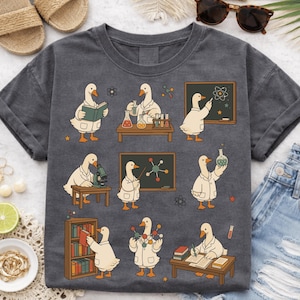 May include: Dark gray t-shirt featuring a whimsical design of cartoon geese in lab coats engaged in various scientific activities. The geese are shown reading, experimenting with beakers, and teaching science. The design includes stars and molecular structures.