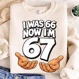 May include: Cream-colored sweatshirt with the text "I WAS 66 NOW I'M 67" in black and white, with the number 67 in large white font. Cartoon hands are drawn below the number. The sweatshirt is folded.