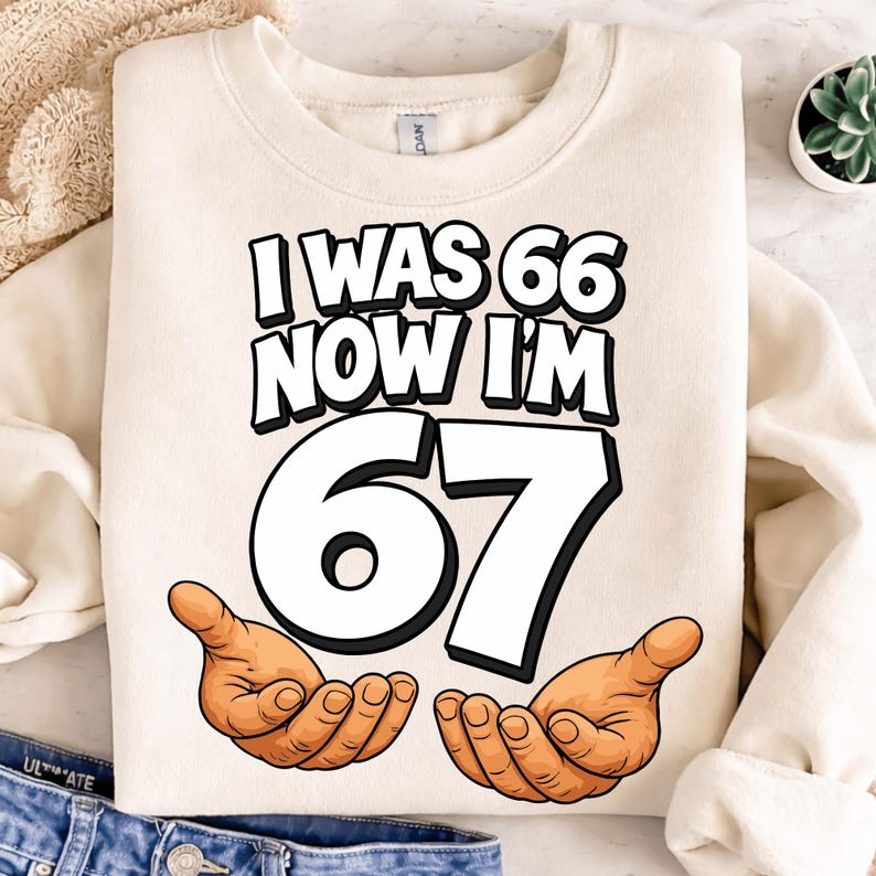 May include: Cream-colored sweatshirt with the text "I WAS 66 NOW I'M 67" in black and white, with the number 67 in large white font. Cartoon hands are drawn below the number. The sweatshirt is folded.