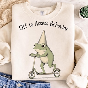 Pode incluir: Camisola creme com o texto "Off to Assess Behavior" acima de um sapo de desenho animado a andar de scooter e a usar um chapéu de festa. O sapo é verde e a scooter é de uma cor verde suave.