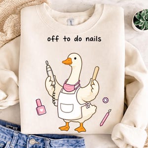 Może przedstawiać: Kremowa bluza z napisem "off to do nails". Grafika przedstawia kreskówkową gęś w różowej maseczce i białym fartuchu, trzymającą wiertarkę do paznokci i drewniany patyczek. Pokazano również lakier do paznokci, klejnot i pilnik do paznokci.