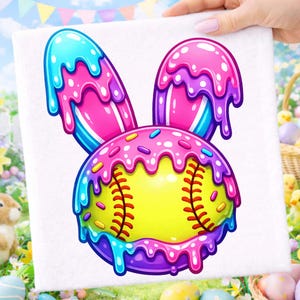 May include: A colorful canvas print featuring a softball designed as a bunny. The softball is yellow with red stitching, and the bunny ears and head are decorated with pink, blue, and purple icing and sprinkles. The canvas is held by a hand.
