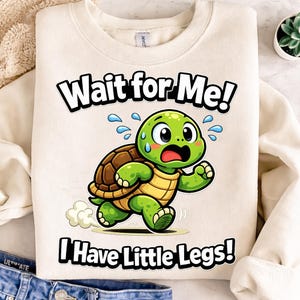 May include: Cream-colored sweatshirt with a cartoon turtle graphic. The green turtle is running with a worried expression, and the text "Wait for Me! I Have Little Legs!" is printed above and below the turtle in black.