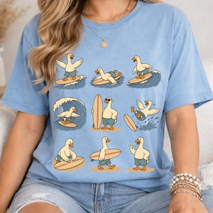 Pode incluir: Uma t-shirt azul-clara com uma estampa gr&aacute;fica de nove patos de desenho animado em v&aacute;rias poses de surf. Os patos usam cal&ccedil;&otilde;es de banho azuis e est&atilde;o representados a surfar ondas, a carregar pranchas e em p&eacute; na areia da praia.