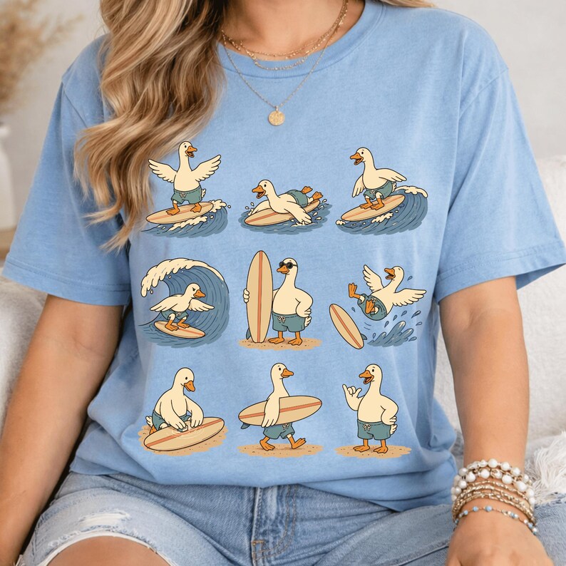 Pode incluir: Uma t-shirt azul-clara com uma estampa gr&aacute;fica de nove patos de desenho animado em v&aacute;rias poses de surf. Os patos usam cal&ccedil;&otilde;es de banho azuis e est&atilde;o representados a surfar ondas, a carregar pranchas e em p&eacute; na areia da praia.