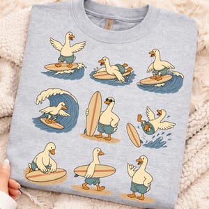 Pode incluir: Uma camisola de gola redonda cinzento-claro com uma estampa gr&aacute;fica repetida de patos brancos a usar cal&ccedil;&otilde;es de banho azuis e a surfar em pranchas de madeira. Os patos est&atilde;o representados em v&aacute;rias poses, a surfar ondas, a carregar pranchas e na areia.
