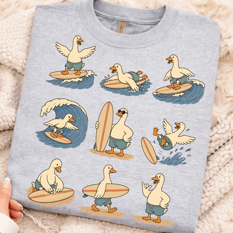 Pode incluir: Uma camisola de gola redonda cinzento-claro com uma estampa gr&aacute;fica repetida de patos brancos a usar cal&ccedil;&otilde;es de banho azuis e a surfar em pranchas de madeira. Os patos est&atilde;o representados em v&aacute;rias poses, a surfar ondas, a carregar pranchas e na areia.