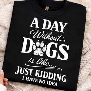 Puede incluir: Sudadera negra con texto blanco: "A Day Without Dogs is like... JUST KIDDING I HAVE NO IDEA". La palabra "Dogs" incluye una huella de pata. La sudadera se muestra sobre una superficie suave y texturizada.